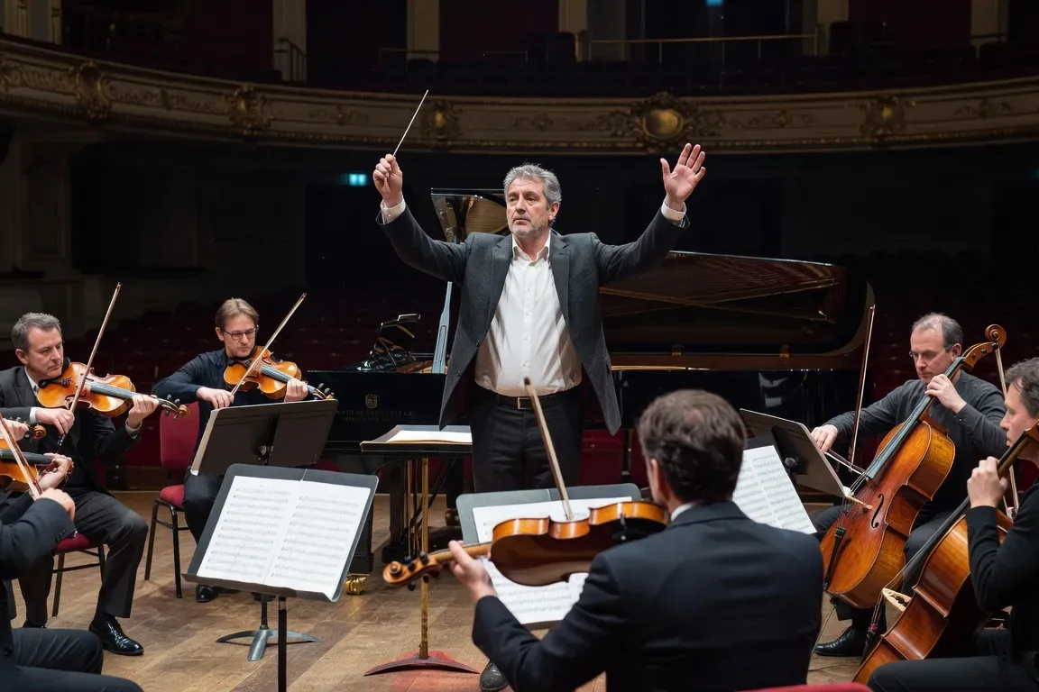 Répétition d'orchestre sur scène de grande salle, chef de face, pianiste au premier plan, cuivres et cordes en arrière-plan